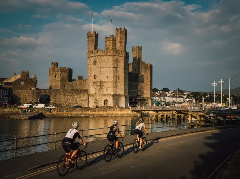 Cyclists going pastt a river next to a grand castle.