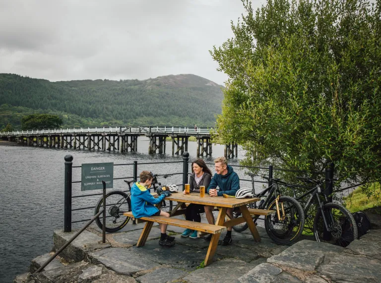 A family sitting on a picnic bench with their bikes, with views of an estuary beyond.