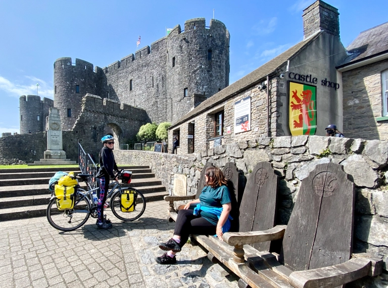A lady on a cycle talking to a woman on a bench by a castle.