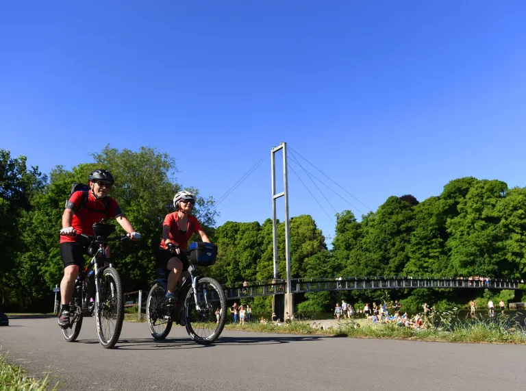 A couple cycling though a park with a footbridge over the river in a distance.