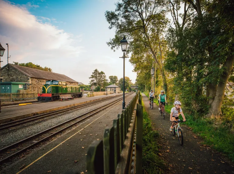 A family cycling past a railway track with a steam train at the station.
