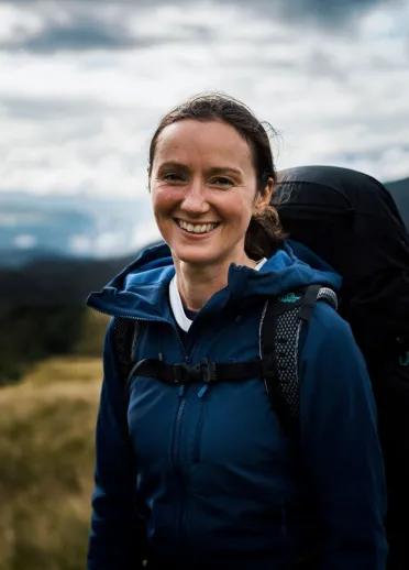 Claire Copeman smiling at the camera wearing outdoor gear and carrying a backpack with mountains in the background.