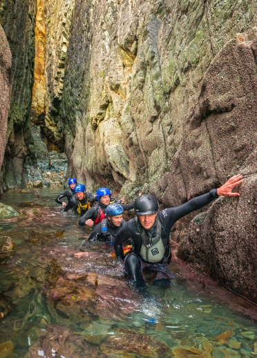 Group of people in a line coasteering in wet suits and helmets.