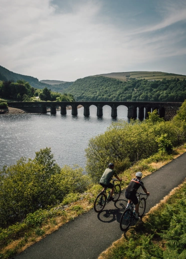 Cyclists on a path near a reservoir heading toward a water bridge.