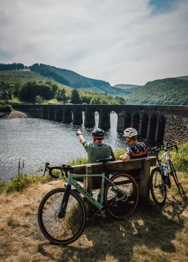 Two cyclists sitting on a bench overlooking a lake and a stone bridge.