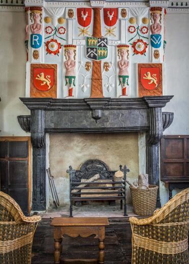 A grand fireplace with wicker chairs in front of it and an ornate medieval decorative cloth above it.