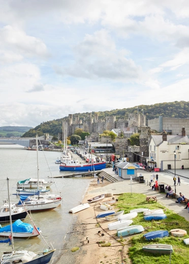 People walking along a harbour with moored boats, and a castle in the background.