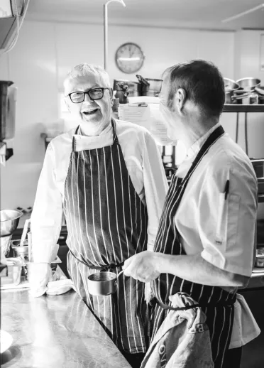Two chefs standing in a restaurant kitchen, laughing.