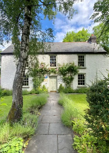 A white cottage at the end of a path with manicured lawns and foliage.