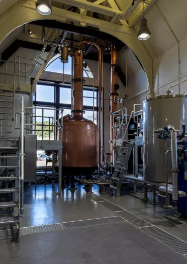 A large copper vat and distilling equipment in a building with large windows.