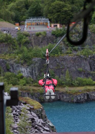 A person flying down on Zip World Velocity zip wire with the blue waters of Penrhyn Quarry beneath.