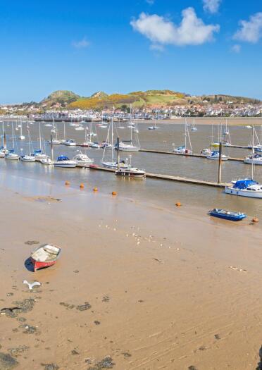 Yachts berthed on the river Conwy alongside a sandy beach and Llandudno Junction in the background.