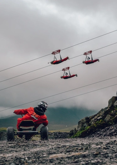 A go carter driving down a quarry with people passing by on zip lines.