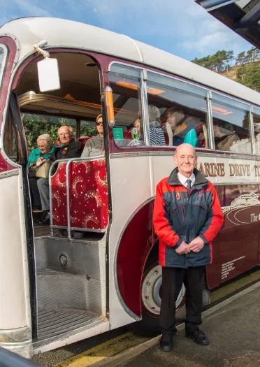 A coach driver standing next to an old fashioned touring coach full of people.