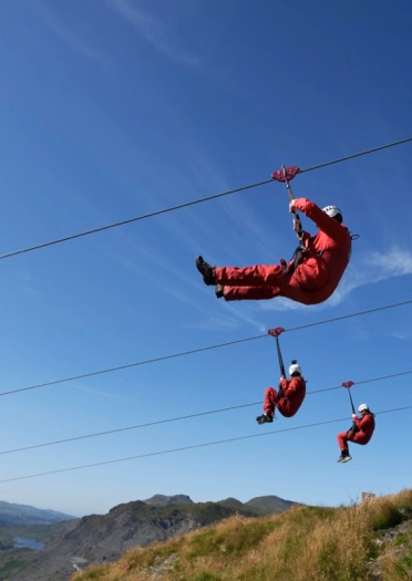 Three people sitting in a harness on parallel zip lines.