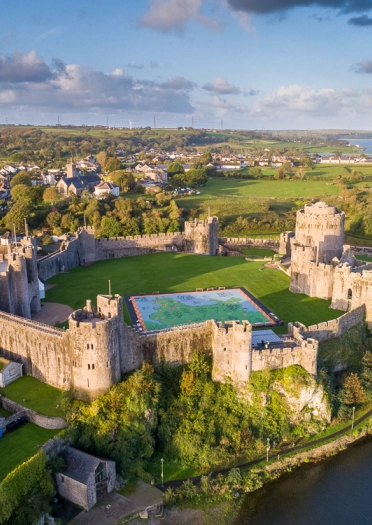 An aerial shot of a large castle with towers alongside the banks of a pond.
