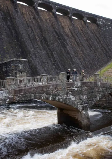 People standing on a bridge over a lake with a large dam in the background.