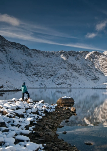 A person looking out at snow covered mountains, reflecting in the lake.