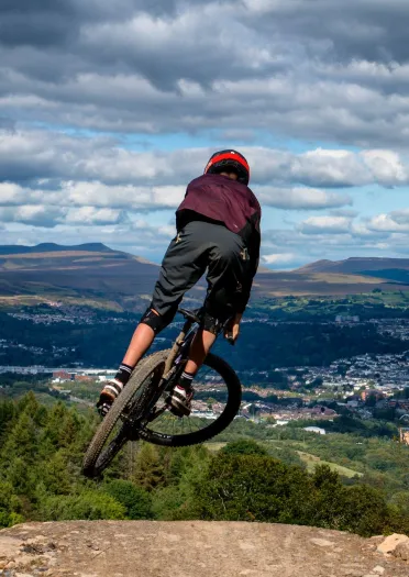 A biker doing a stunt jump at the top of a mountain with views beyond.