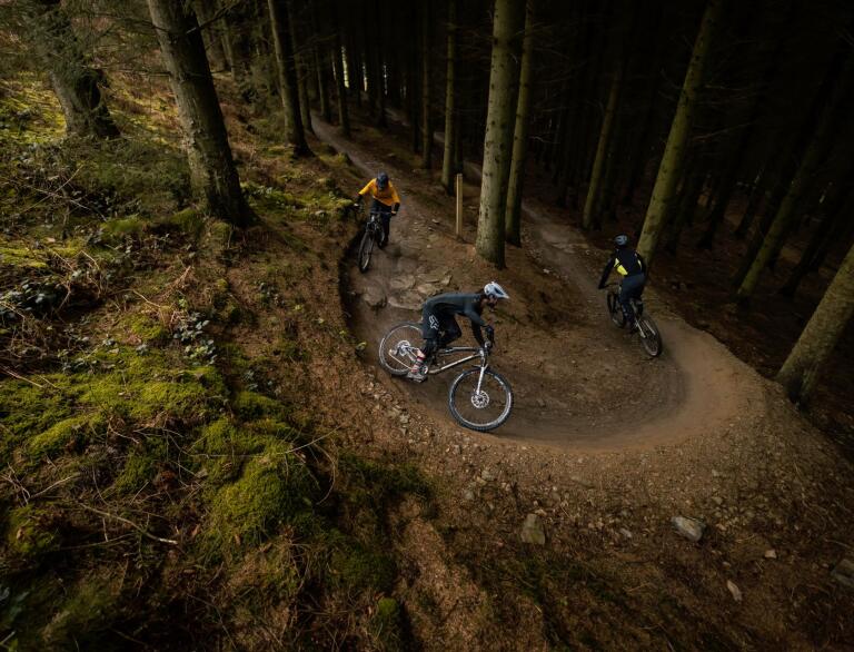 Three mountain bikers going around a curved dirt track in a forest.