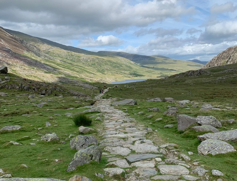 Walkers on a stone path leading to a lake surrounded by mountains.