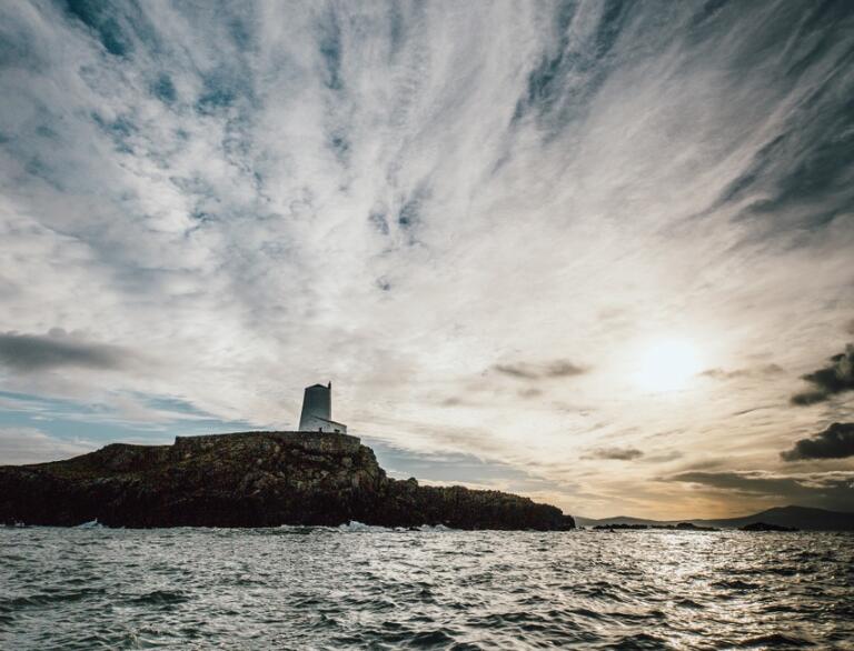A shadowy site of and island with a lighthouse with waterfall clouds in the sunset.