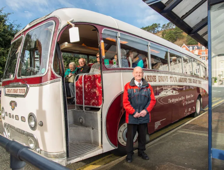 A coach driver standing next to an old fashioned touring coach full of people.