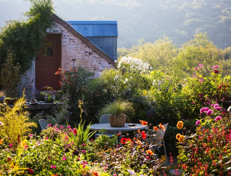 A garden building surrounded by flora and fauna and a garden table and chairs.