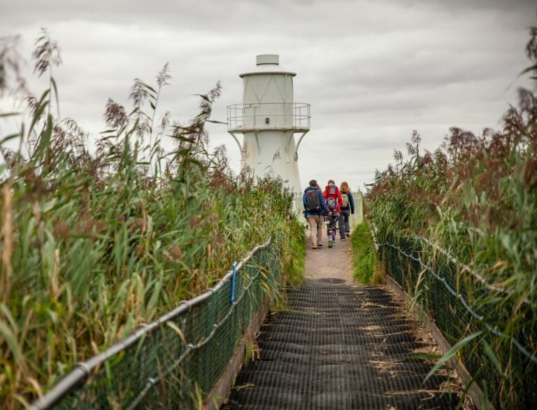 Group of people walking on a boardwalk toward a lighthouse.