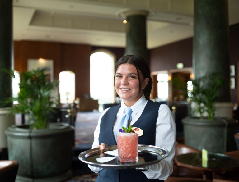 A waitress smiling at the camera with a cocktail on a serving tray.