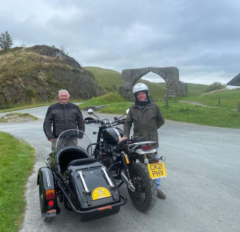 Two gentlemen standing my a motorcycle and sidecar with hillside views beyond.