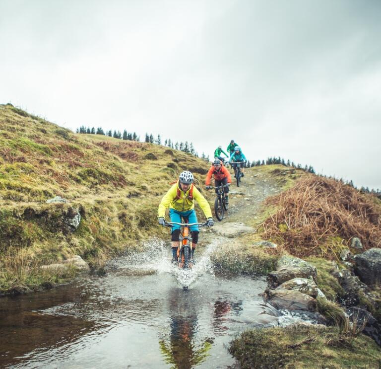 Three mountain bikers cycling downhill through a puddle on a purpose built dirt track.