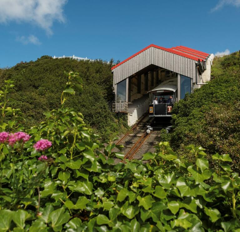A mountain cliff railway departing from it's shed framed by foliage and flowers.