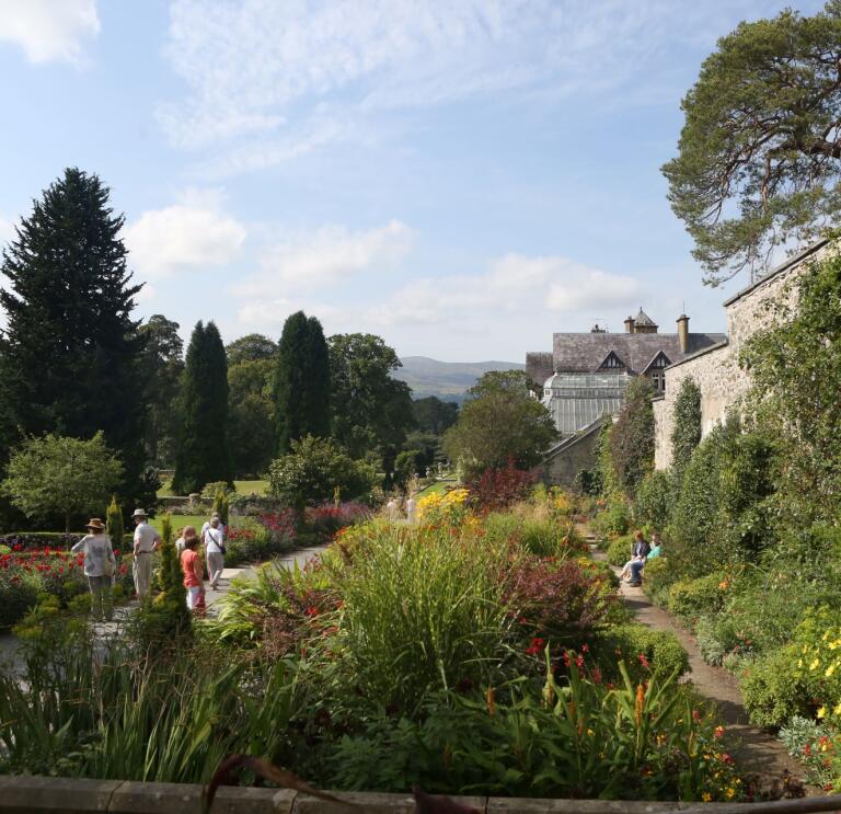 Flowers, trees and shrubs alongside paths and stone walls with a grand house in the background.