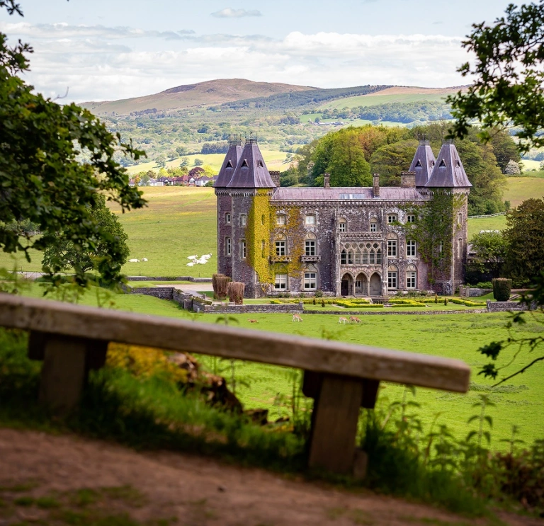 A large manor house with its towers covered in creeping ivy viewed from a seating bench.