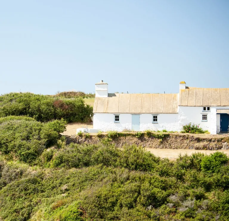 A walker on a coastal path leading to a white cottage.