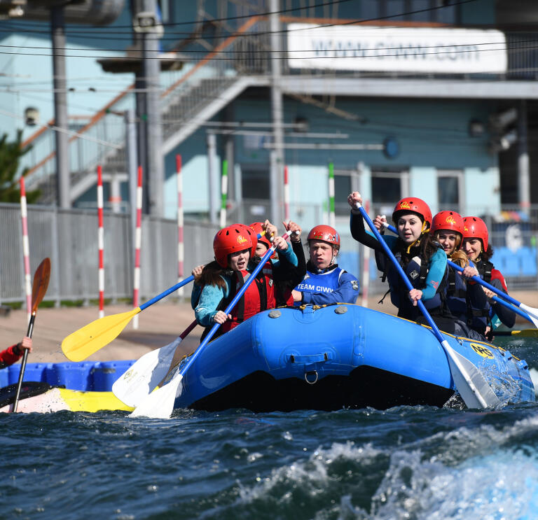 A group kitted out in helmets and lifej ackets using their oars to take on the rapids in a blue dinghy.