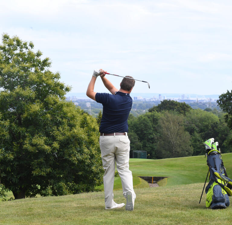 Golfer having taken a swing next to his golf bag and fellow player with view of Cardiff beyond.
