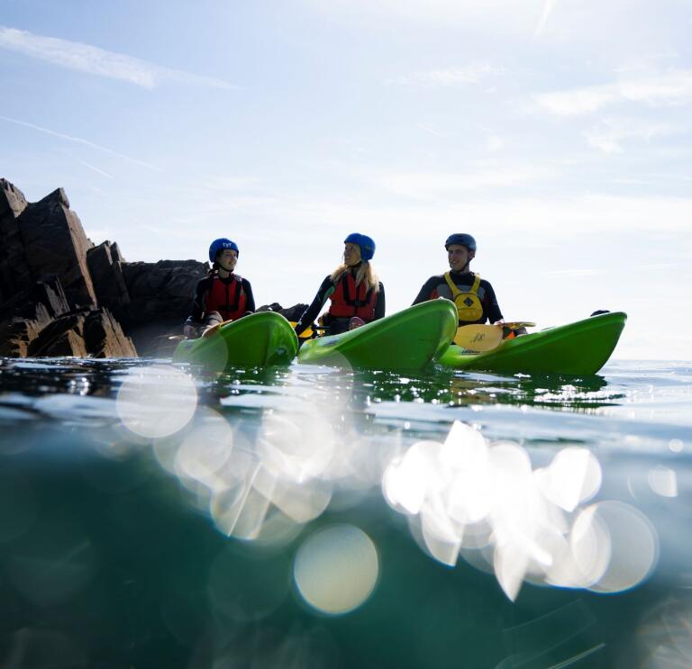 Three kayakers on the ocean with a rocky cove backdrop.