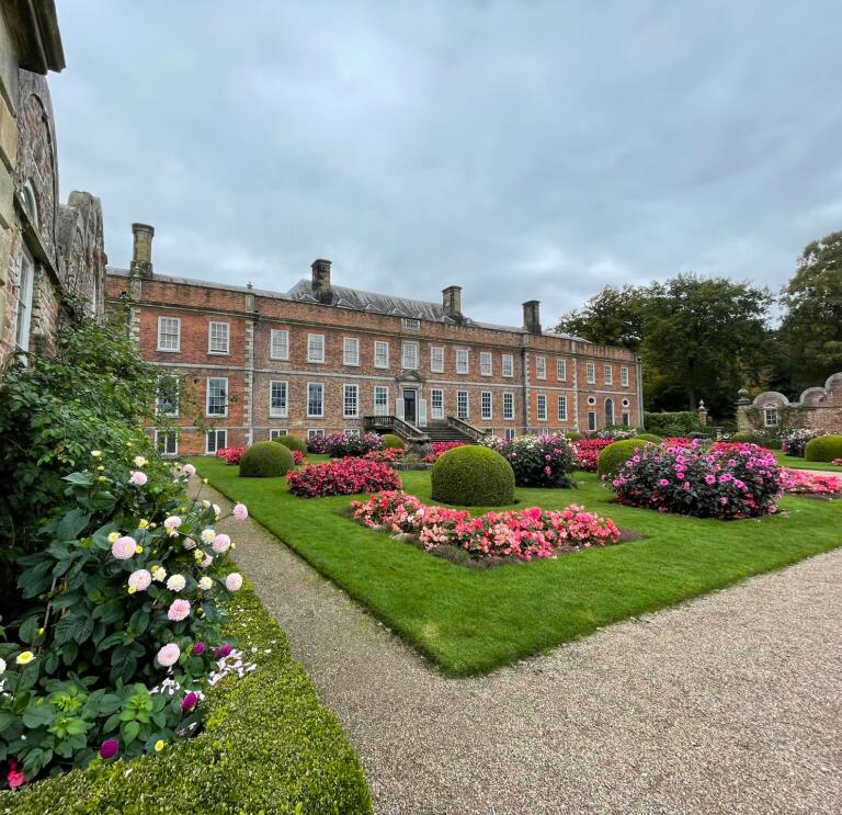 A grand house with beautiful flowers and round shrubbery in the forecourt.