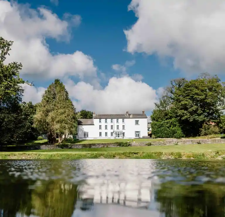 A white manor surrounded by trees reflecting in a lake.