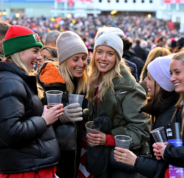 Group of ladies holding drinks and laughing, outside a sports stadium .