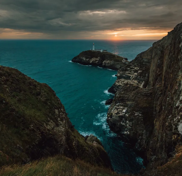 Person standing on a coast path overlooking a lighthouse at sunset.