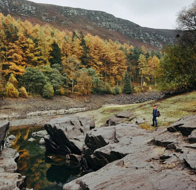 A walker by a lake surronded by rocks and an autumnal forest.