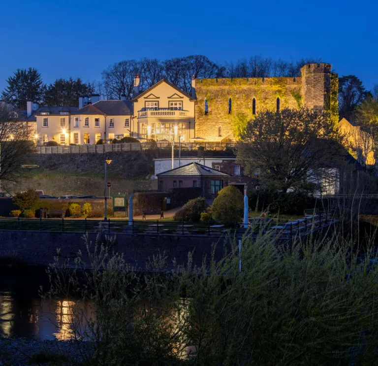 External shot of a castle hotel lit up at night, with a river running beside it.