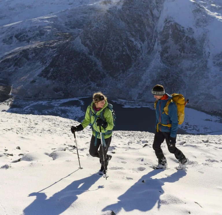 A couple walking on a snowy mountain with a frozen lake below.