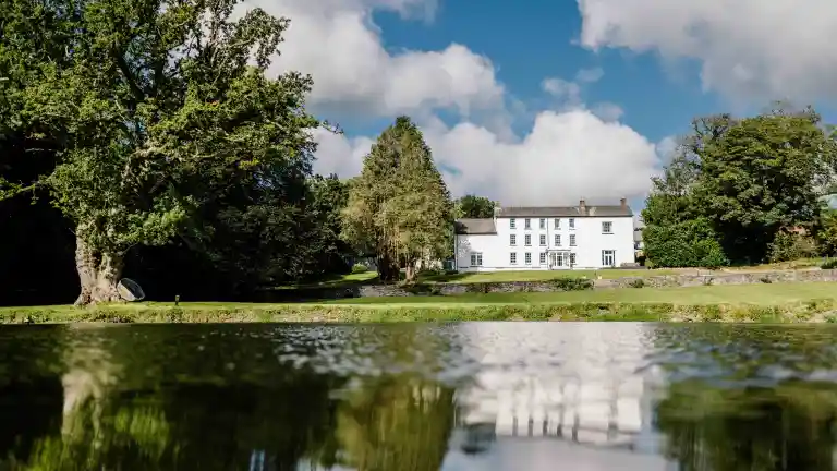 A white manor surrounded by trees reflecting in a lake.