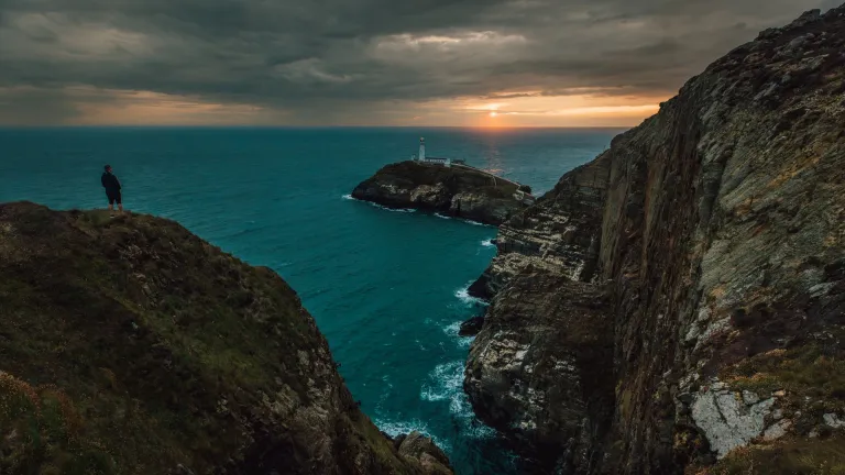 Person standing on a coast path overlooking a lighthouse at sunset.