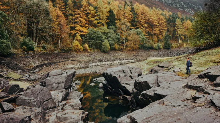 A walker by a lake surronded by rocks and an autumnal forest.