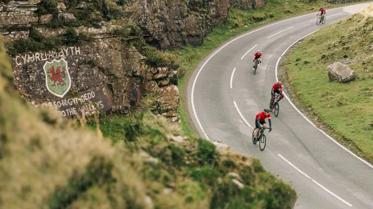 Cyclists on a road against a mountainous landscape with a Welsh dragon on the wall.